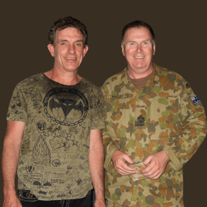 Cold Chisel's Ian Moss with WO1 Mark Langley in East Timor. Ian Moss is wearing a green t-short with black detail and Mark is wearing his Army camouflage. They are standing close together and smiling at the camera.