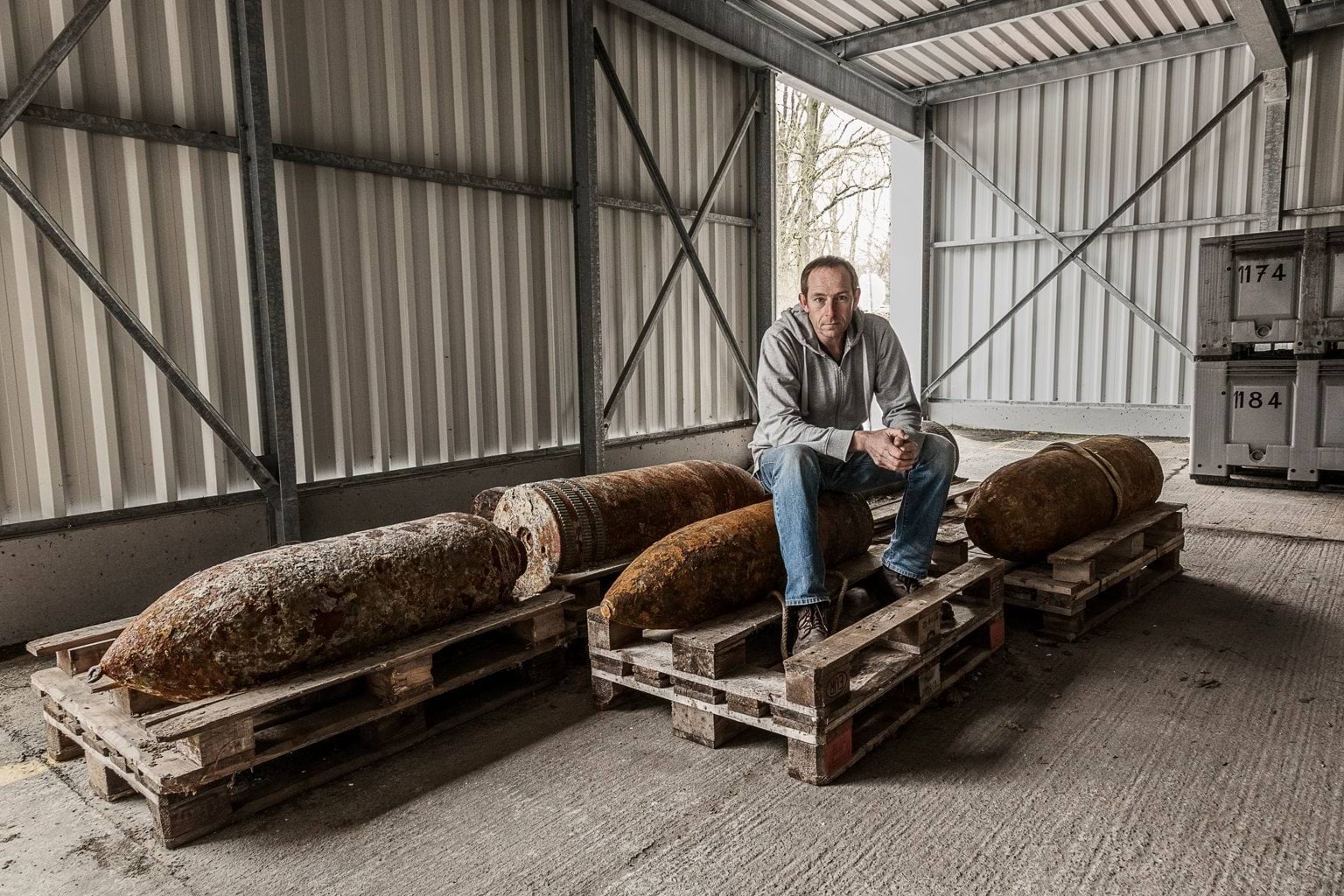 A man in a grey jumper and blue jeans sitting on recovered ammunition from the First World War. He is staring straight into the camera with a solemn look on his face. Around him are three other large shells on wooden crates.