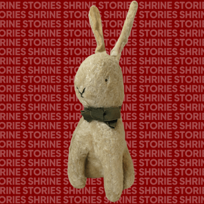 the image, a stuffed rabbit is shown against a red background patterned with the repeated text "SHRINE STORIES" in bold, uppercase letters. The rabbit has a simple, hand-stitched face, long ears, and wears a slightly frayed green bow around its neck.