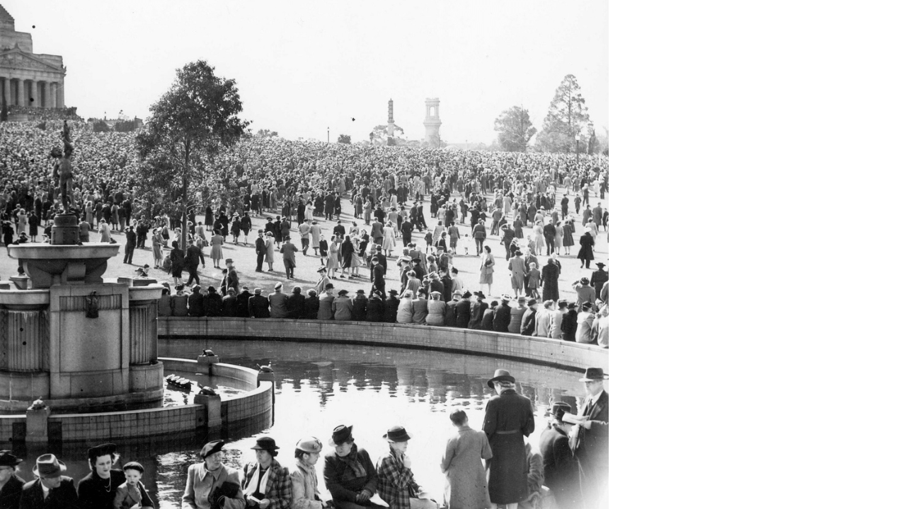 Black and white image of crowds gathered at Melbourne’s Shrine of Remembrance after the declaration that the Second World War was over in 1945. Some people sit around a fountain, while a large gather on the grass in front of the memorial.