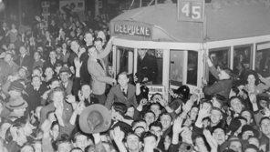 A black and white image displaying a large crowd of people surrounding and climbing on a tram in Melbourne as they celebrate the Victory in the Pacific, 1945.