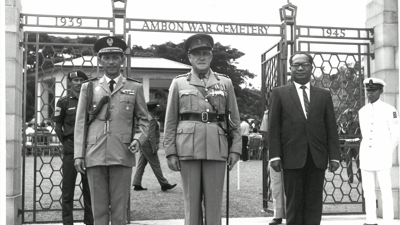 Black and White of 3 men standing in front of the Ambon War Cemetery, other people in the background.