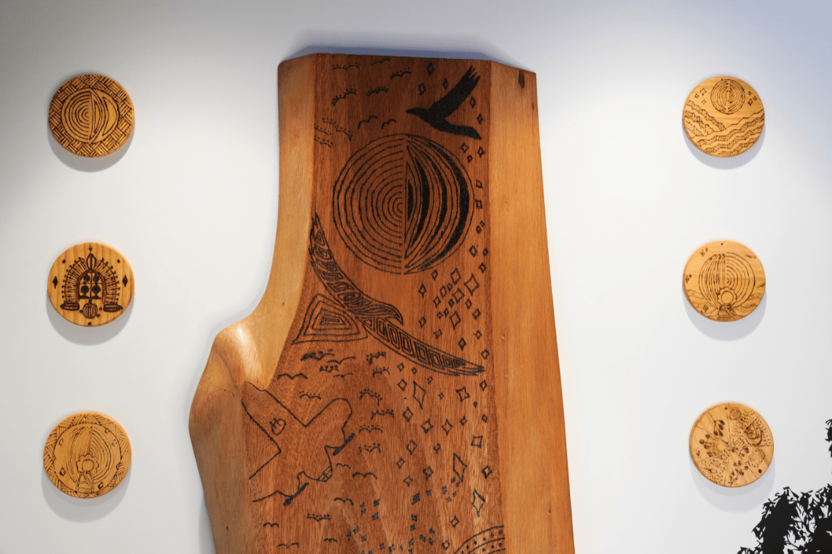 A wooden board with black pyrographic art depicting a bird in flight, a circular motif divided in half, and abstract shapes resembling waves and stars.