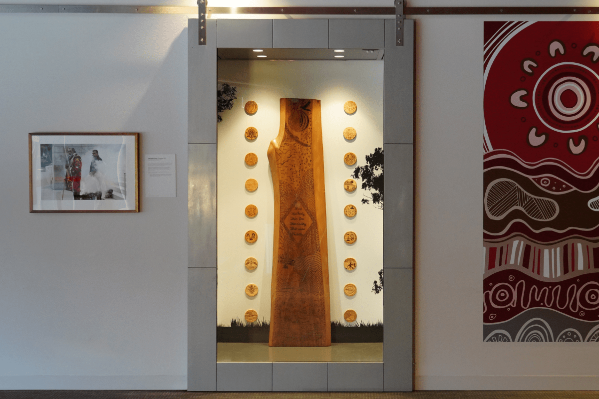 A large, vertical piece of wood with carvings is displayed in a glass case.